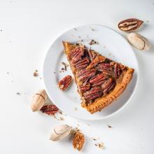 a slice of pecan pie on a white plate on a white surface with pecans and pecan pieces scattered around it