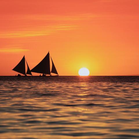 two sailboat silhouettes against a red sky at sunset with the sun just dipping below the horizon of the ocean