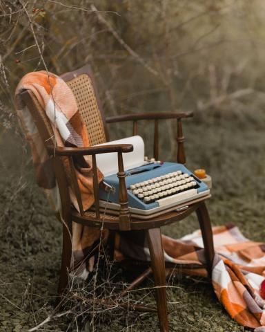 outside shot of a wooden framed chair with orange and white cushions on which rests a vintage typewriter via Canva