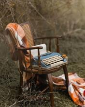 outside shot of a wooden framed chair with orange and white cushions on which rests a vintage typewriter via Canva